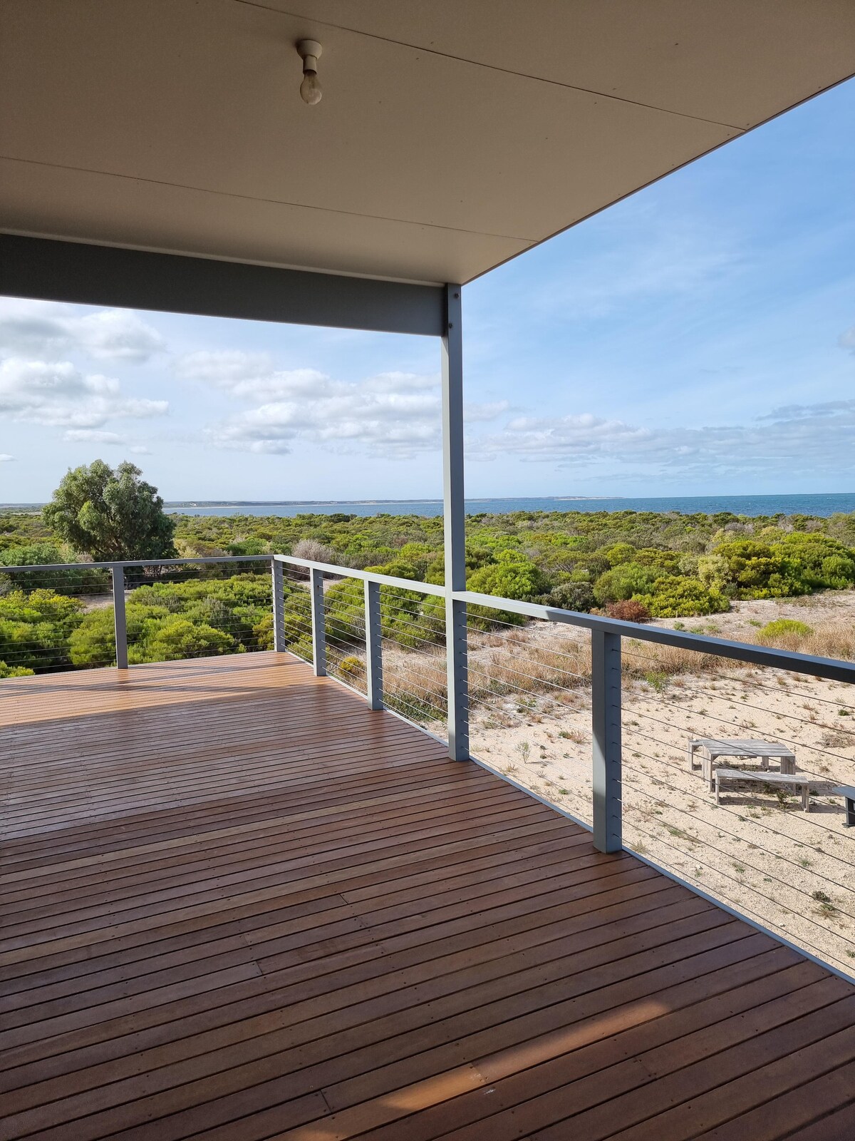 An expansive wooden deck extends outward, offering unobstructed views of lush greenery and the sea in Marion Bay. The clear sky and scattered clouds fill the background, creating a serene atmosphere. The railings are sleek, providing safety while enjoying the outdoor space.