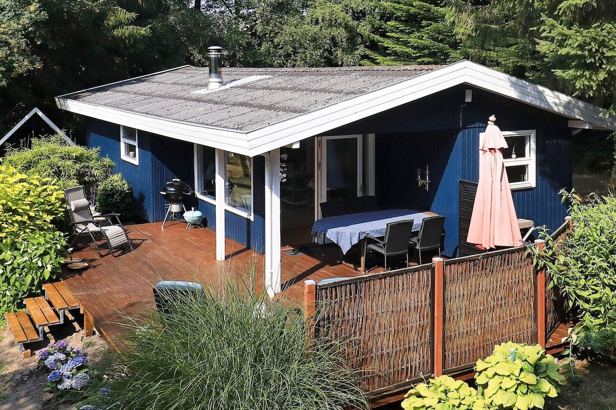 A traditional blue holiday home is situated on a natural plot surrounded by greenery. An outdoor terrace with a dining table and chairs is featured, accompanied by a parasol. The property includes a small BBQ area and is surrounded by vibrant plants and shrubs.