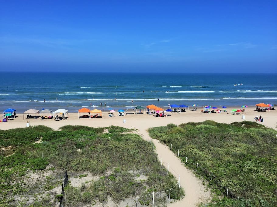 A sandy beach stretches under a bright blue sky, where colorful tents and umbrellas provide shade for beachgoers. Gentle waves lap at the shore, and patches of grass line the sandy path leading to the water, offering a natural transition from land to sea.