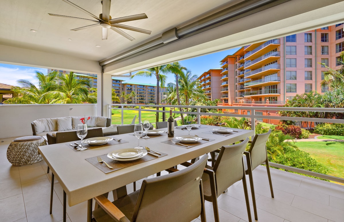 An expansive dining area on the balcony is furnished with a large table set for six, complemented by comfortable chairs. The view includes lush greenery and nearby resort buildings, framed by palm trees under a clear blue sky.