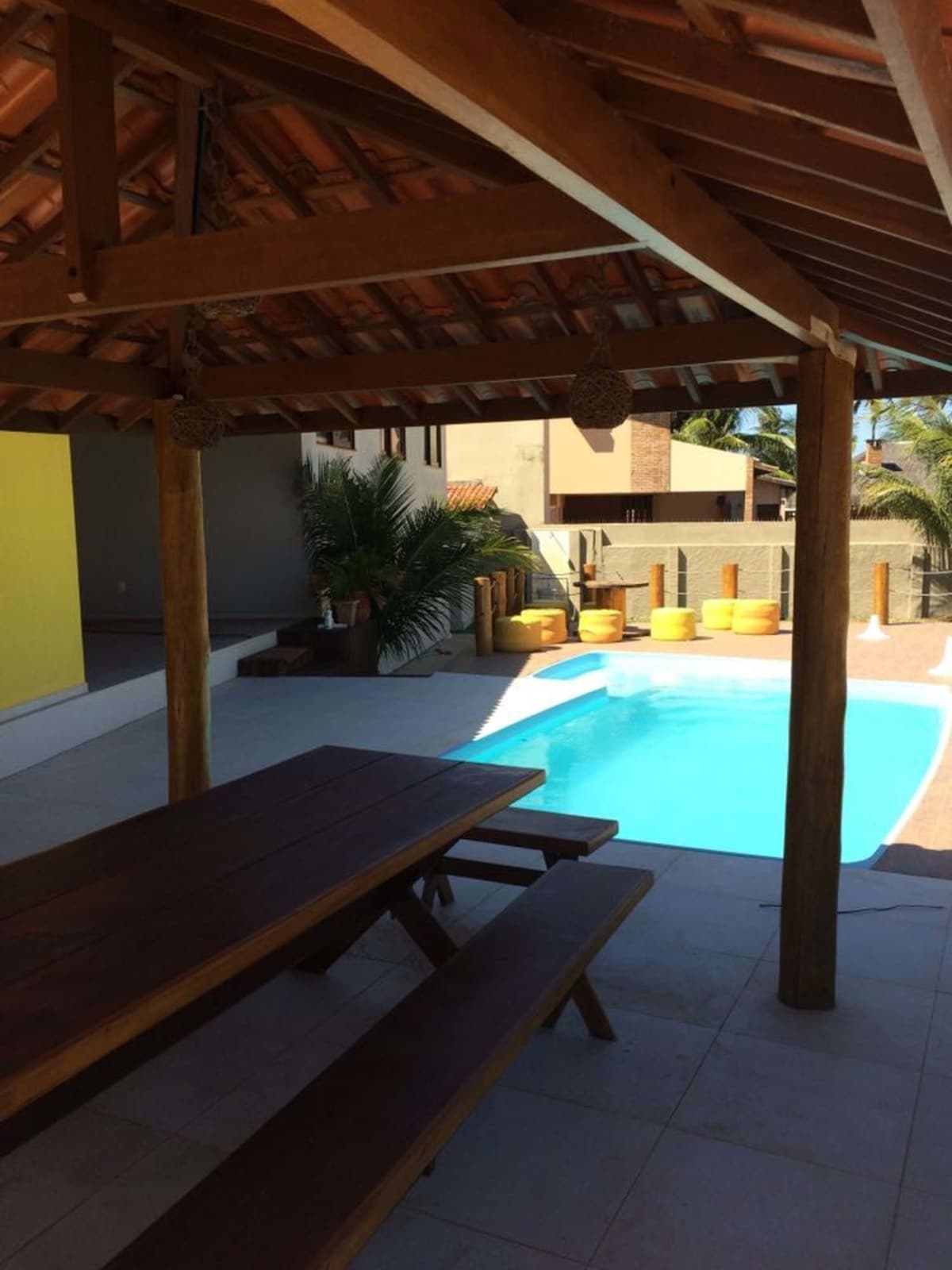 A shaded outdoor area features a wooden table set with benches, overlooking a refreshing swimming pool. Yellow lounge chairs line the poolside, providing spots for relaxation. Lush greenery is visible nearby, enhancing the inviting space for alfresco dining or lounging.