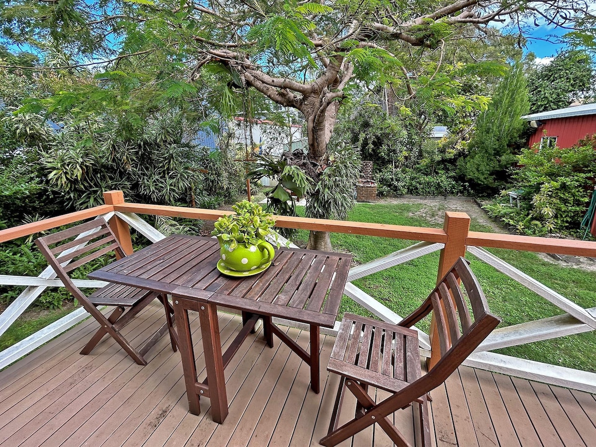 An outdoor dining area is set upon a wooden deck, featuring a table surrounded by two slatted chairs. A green planter with foliage sits at the center of the table. Lush greenery is visible in the background, with trees and grass creating a serene outdoor space.