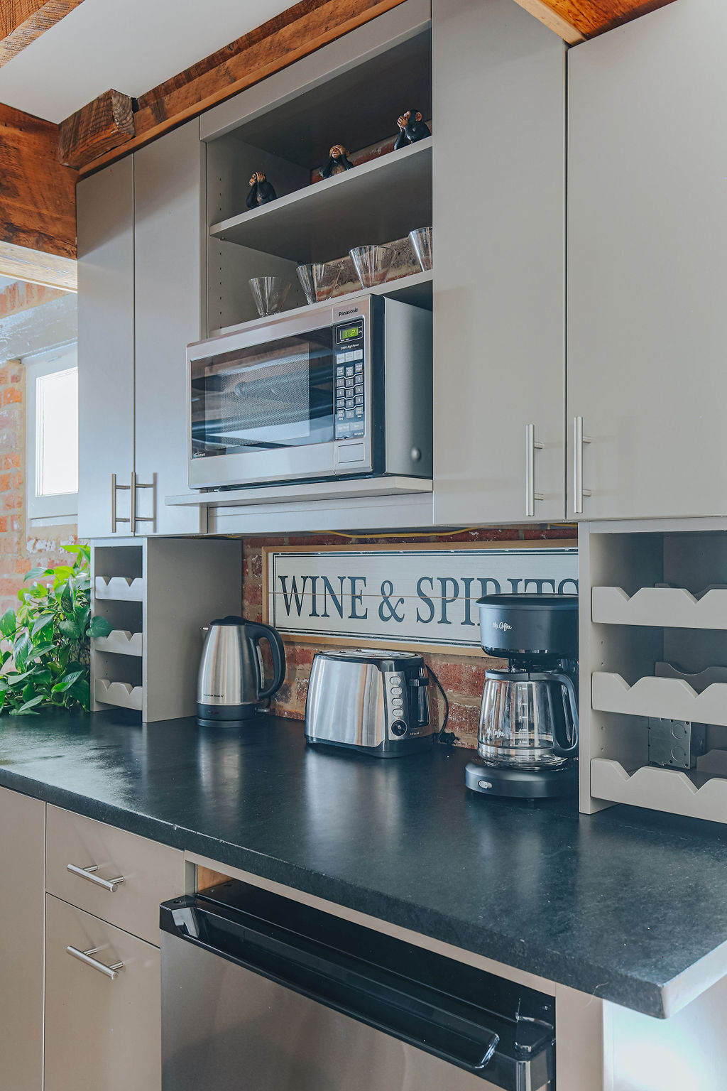 A modern kitchenette features sleek cabinetry with an open shelf displaying glassware. A microwave is positioned above a countertop that includes multiple appliances: two electric kettles, a coffee maker, and a mini fridge. Decorative shelving for wine storage is also visible.