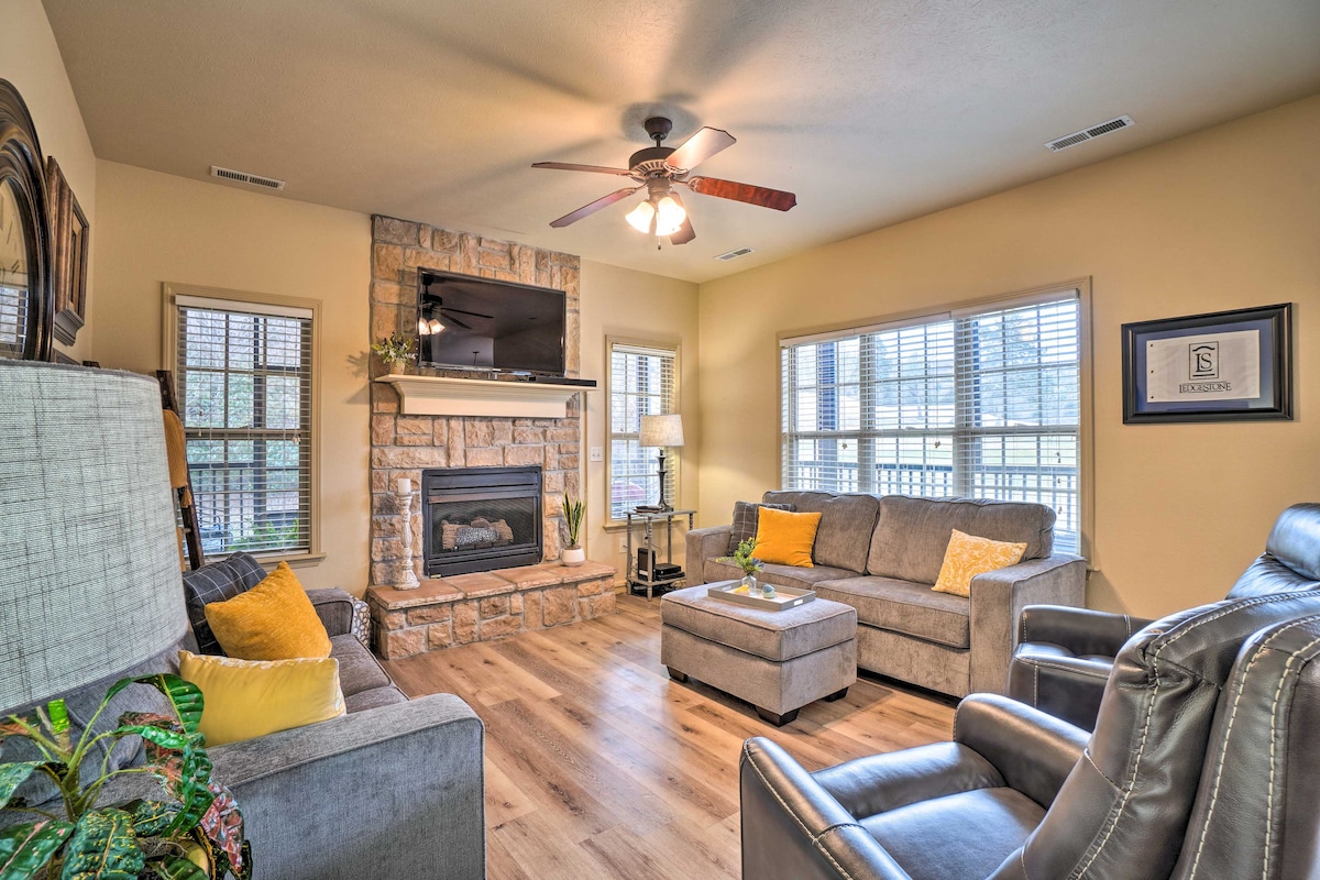 A comfortable living area is highlighted by a stone fireplace and a ceiling fan. Soft gray sofas and a recliner are arranged around a coffee table. Natural light filters through the window blinds, creating a warm and inviting space.