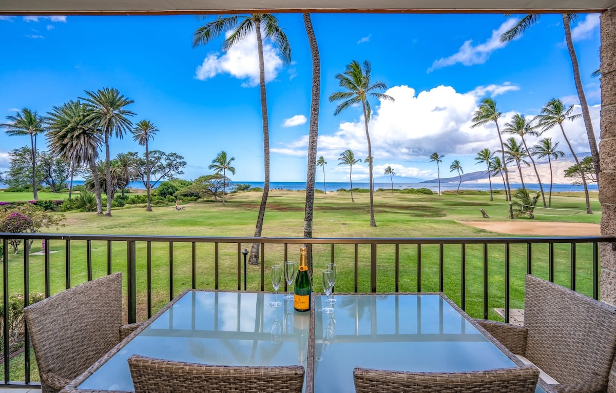A spacious lanai features a glass-top dining table surrounded by woven chairs. The view showcases swaying palm trees and lush greenery, leading to an ocean vista under a bright blue sky with scattered clouds.