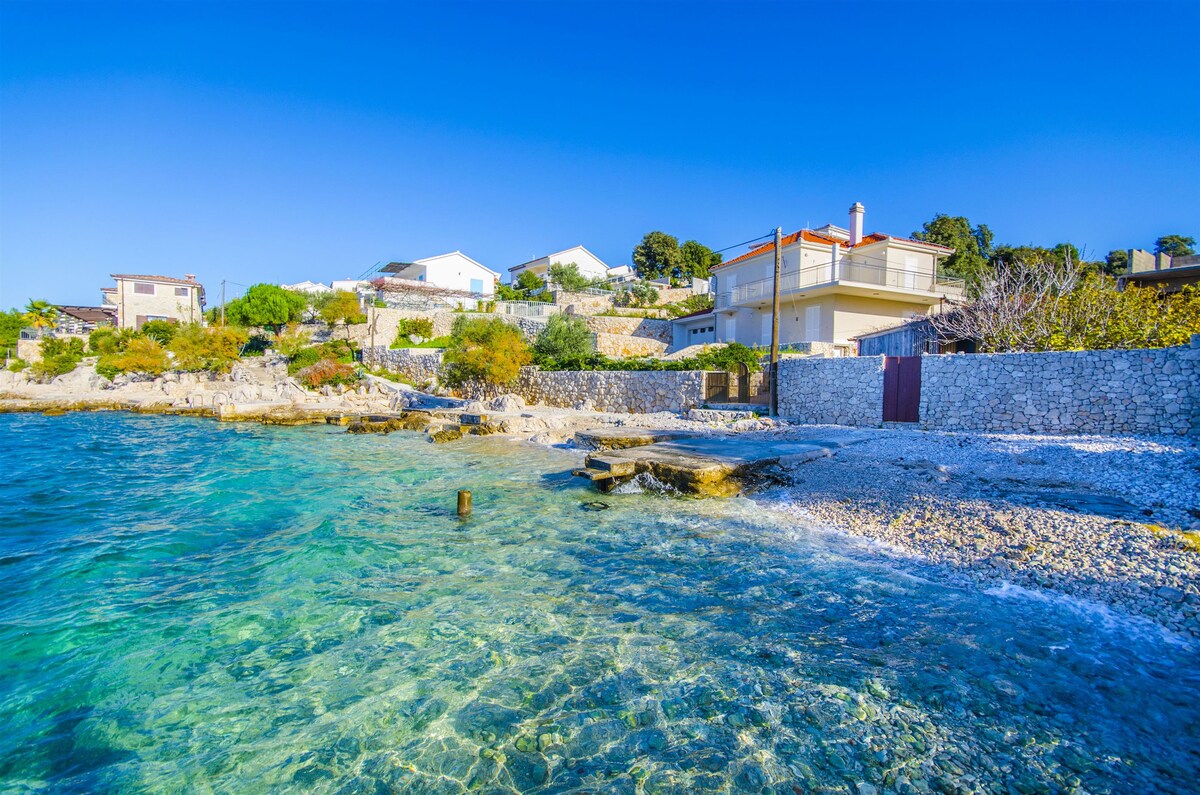 The image captures a serene coastal scene with clear turquoise water lapping against a rocky shore. Houses are visible on the hillside, surrounded by greenery. The natural landscape features smooth stones and gentle waves, contributing to a calming seaside atmosphere.