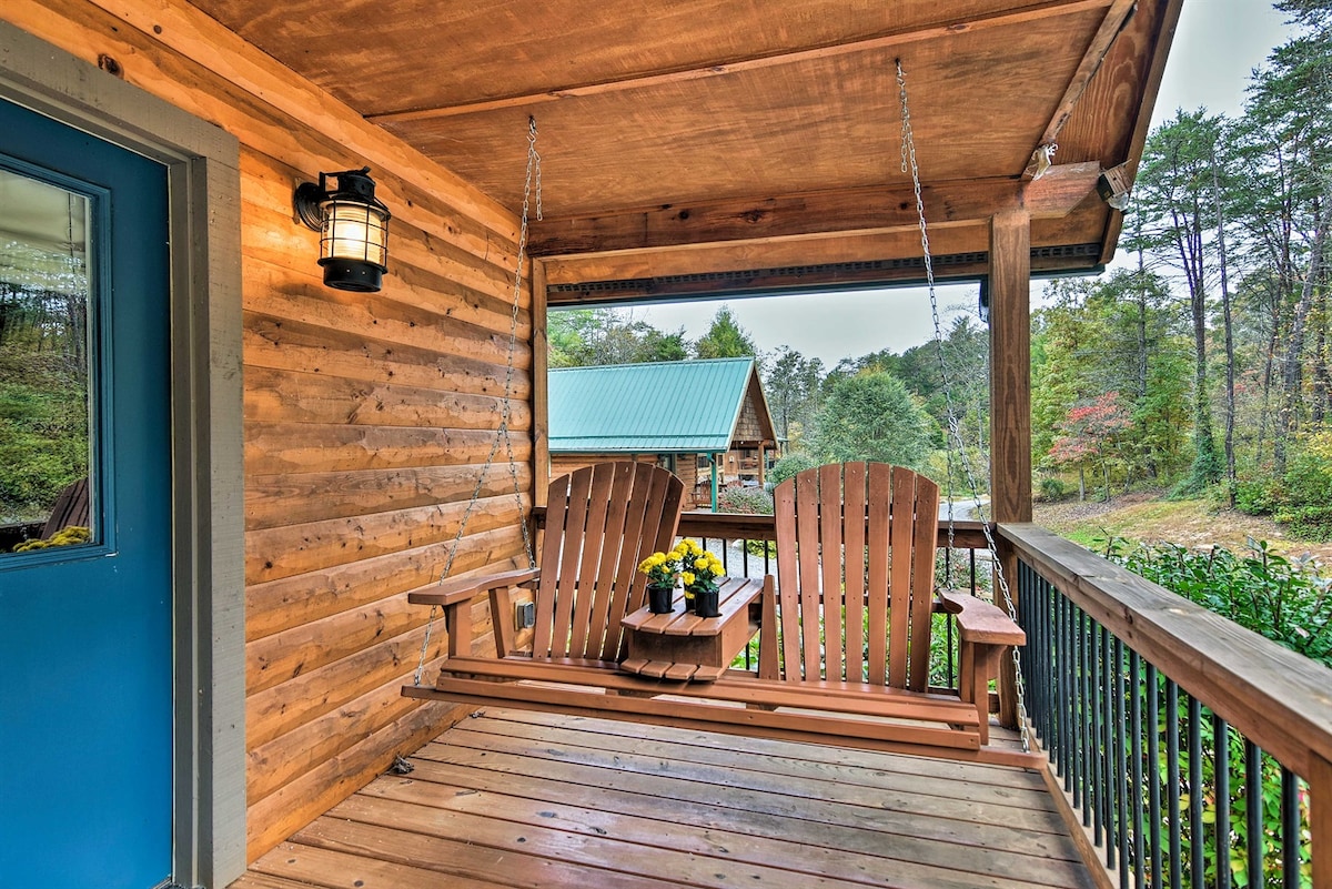 A covered porch features a swinging bench with two seats, adorned with a small table holding a potted flower arrangement. Wooden siding and a blue door complement the natural surroundings, with glimpses of foliage and distant cabins visible in the background.