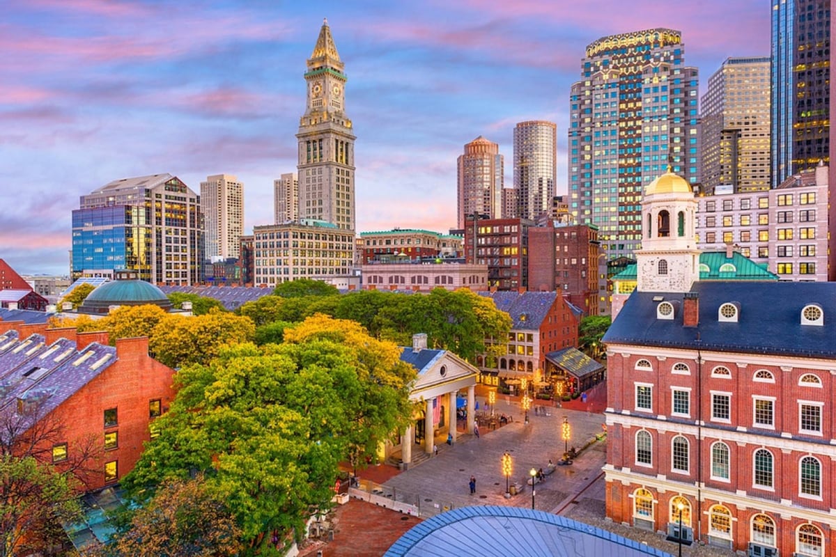 The vibrant city skyline of Boston is displayed, featuring a mix of historic and modern architecture. The tall clock tower stands prominently among newer buildings, while lush greenery fills the foreground. A colorful sky at dusk enhances the scene, reflecting the lively ambiance of the area.