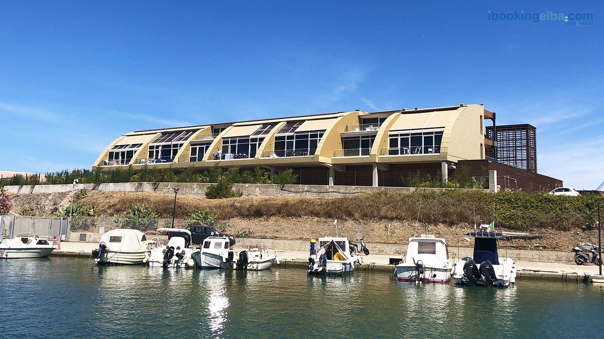 A modern two-level building is located near a serene marina, overlooking a row of small boats. Large windows are visible along the upper floor, offering a glimpse of views and natural light. The surrounding area features greenery and well-maintained paths.