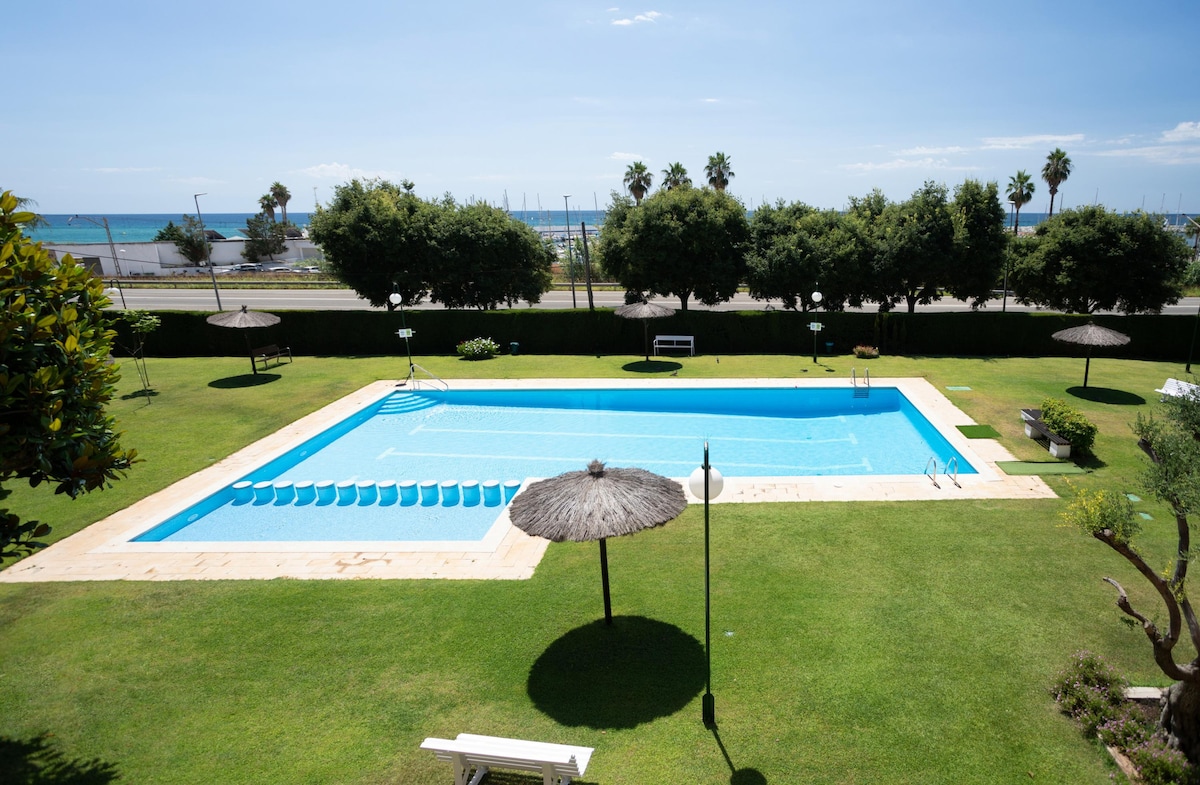 A spacious swimming pool is set within a well-maintained grassy area, surrounded by palm trees and umbrellas for shade. Clear blue water glistens under the sun, while the nearby beach and marina are visible in the background.