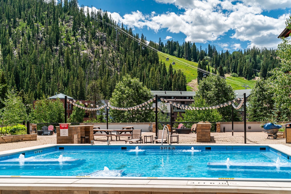An outdoor swimming pool is visible, flanked by landscaped greenery and picnic areas. Nearby, chairlifts ascend the lush hillside. The scene is illuminated by natural sunlight, creating a refreshing atmosphere for relaxation or recreation.