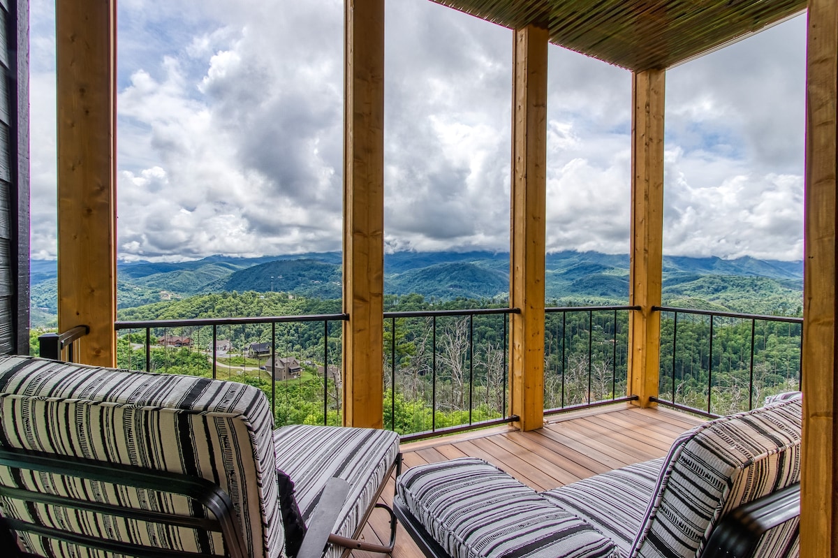 A spacious deck is framed by wooden pillars, providing unobstructed views of the surrounding Smoky Mountains. Two lounge chairs are decorated with striped fabric, inviting relaxation while taking in the expansive landscape under a partly cloudy sky.