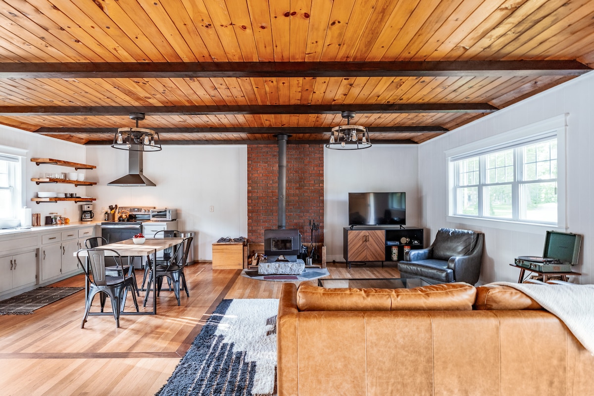 An open living and dining area showcases mid-century modern design with wood beams and a stone fireplace. A comfortable leather sofa faces a TV, while a dining table for six sits adjacent to a well-equipped kitchen featuring open shelving, providing a welcoming communal space.