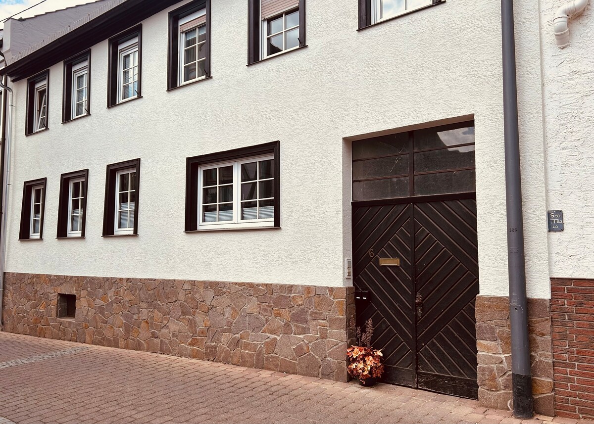 The exterior of a multi-story building is presented, featuring a combination of white stucco and stone elements. Several rectangular windows are positioned symmetrically above a black double door, which displays a diagonal wood pattern. A simple floral arrangement is placed near the entrance.