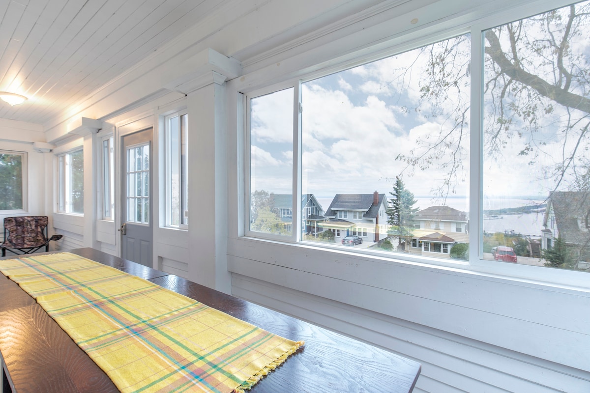 A well-lit sunroom features large windows providing views of Lake Superior and neighboring homes. A long wooden table is set with a yellow and green plaid table runner, creating a cozy space bathed in natural light.