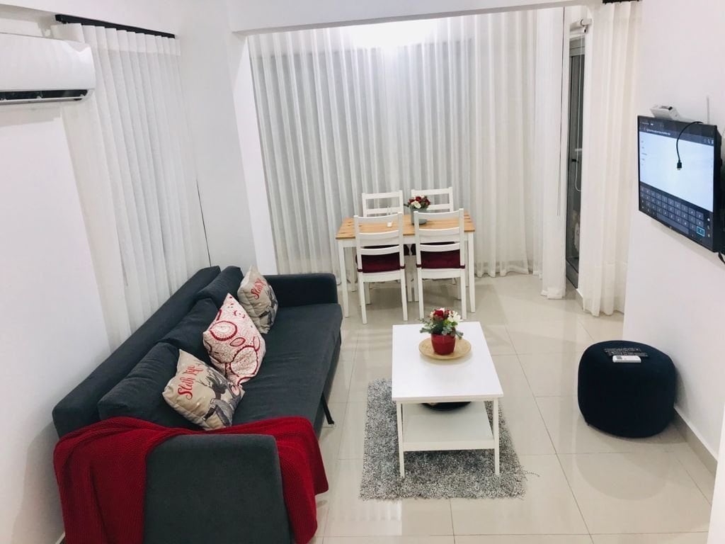 A modern living area features a dark gray sofa adorned with decorative pillows and a red throw. A white coffee table sits atop a soft rug, while a small dining table with four chairs is positioned nearby. Natural light filters through sheer curtains.