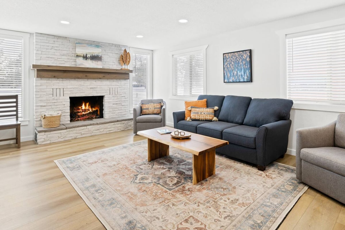 A bright living room features a stone fireplace as a focal point, surrounded by large windows allowing natural light. Comfortable seating includes a dark sofa and two chairs arranged around a central wooden coffee table. A neutral area rug adds warmth to the space.