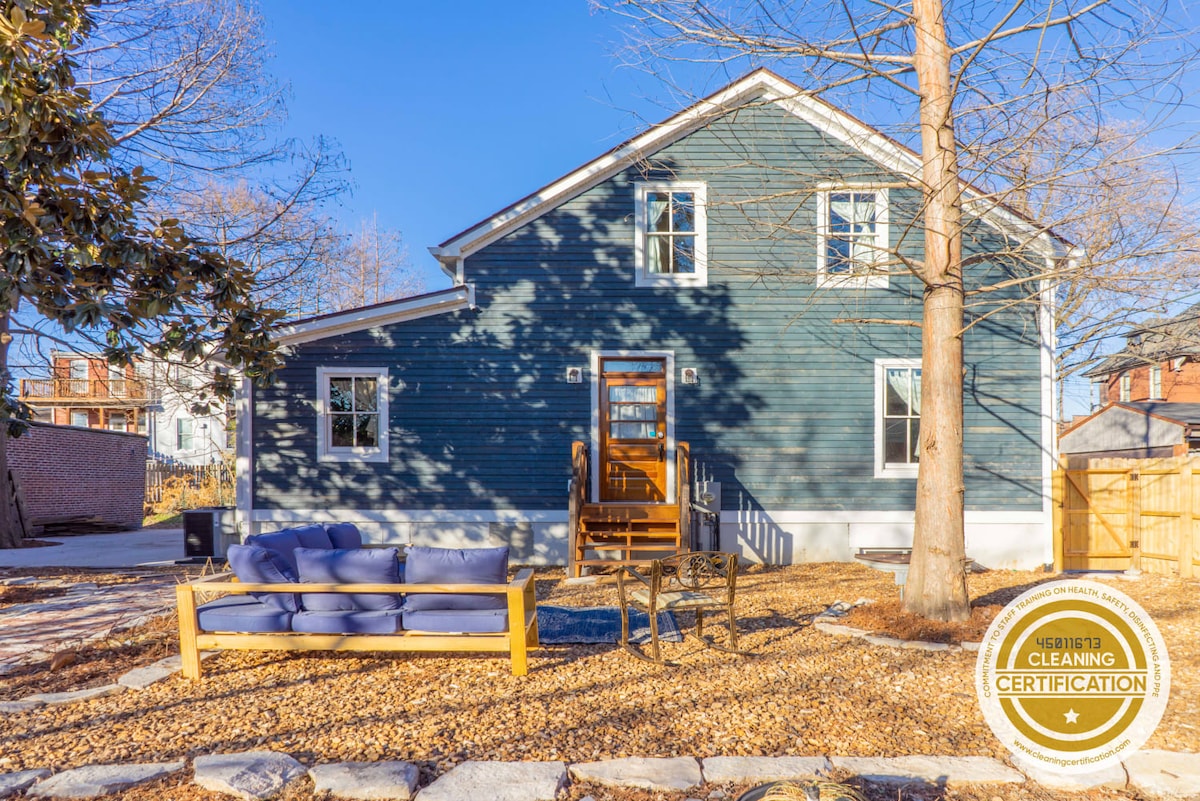 The exterior of a renovated farmhouse is shown, featuring a blue façade with white trim. A comfortable outdoor seating area is placed on a gravel patio, surrounded by trees. Sunlight casts shadows across the facade, highlighting the inviting entrance.