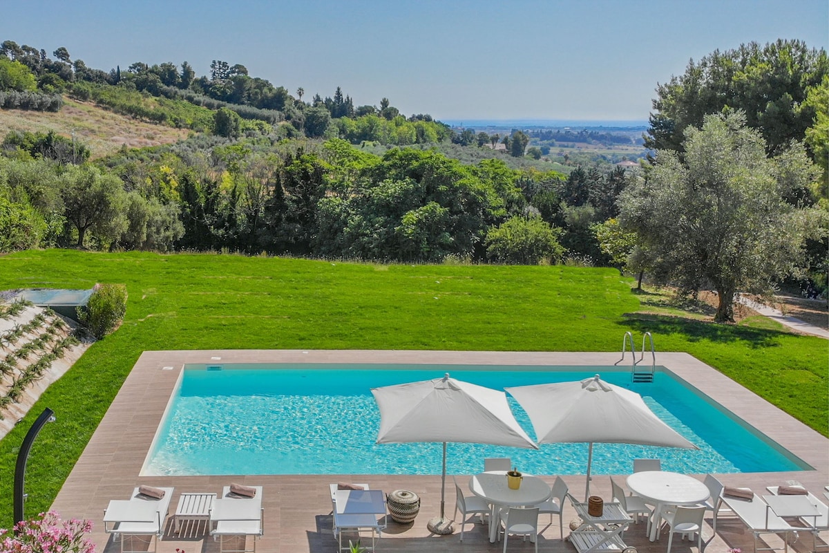 An outdoor space is highlighted by a swimming pool surrounded by green grass and shrubs. Beach umbrellas offer shade over lounge chairs. In the background, rolling hills and olive trees are visible, with a distant view of the sea adding to the serene setting.