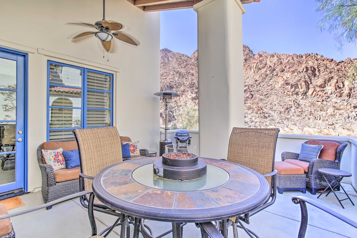 A furnished patio area features a round dining table with a central fire pit. Comfortable seating includes cushioned chairs and a barbecue grill nearby. The backdrop showcases rugged mountain views under a clear blue sky, and a ceiling fan hangs overhead, adding to the outdoor ambiance.