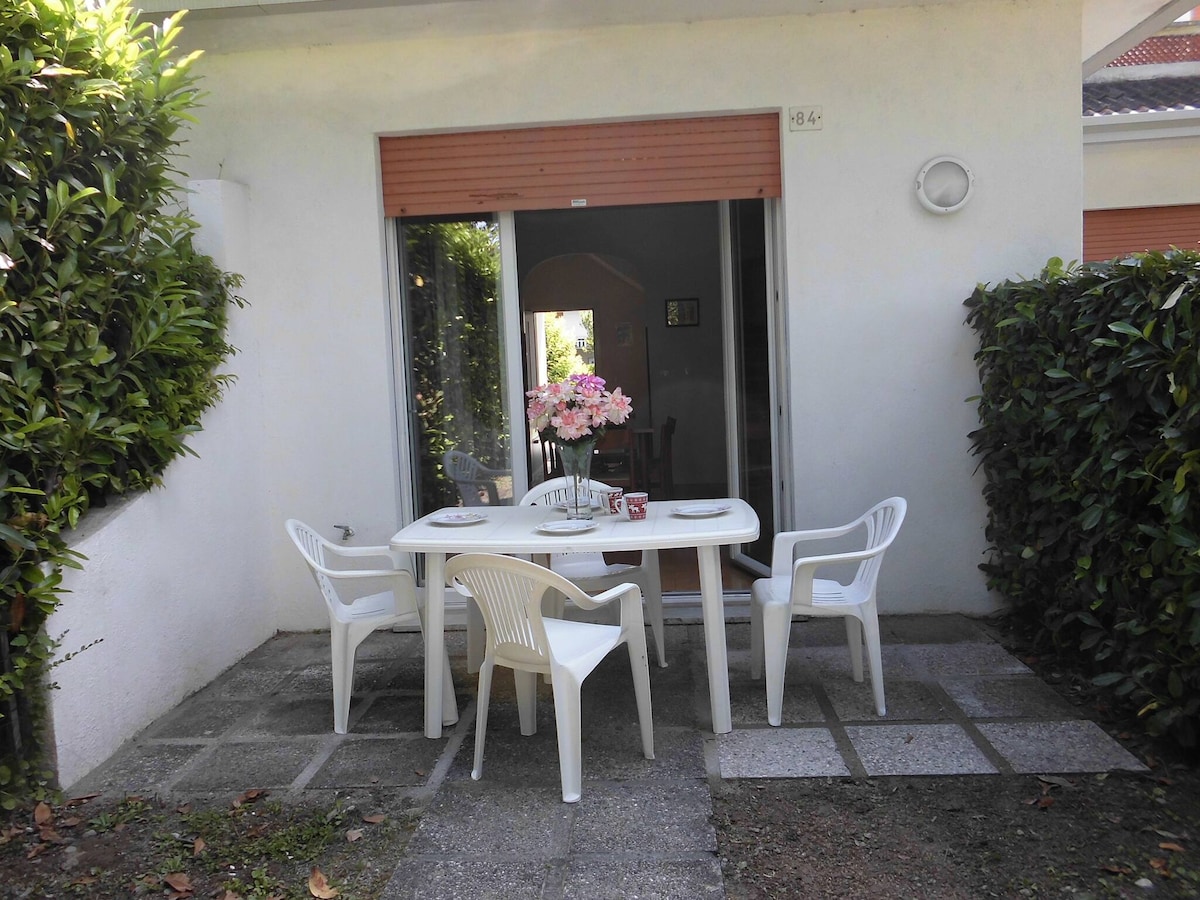 An outdoor seating area is showcased, featuring a table set for four. White plastic chairs surround the table, which is adorned with a floral centerpiece. Lush green hedges flank the space, providing privacy and natural ambiance.
