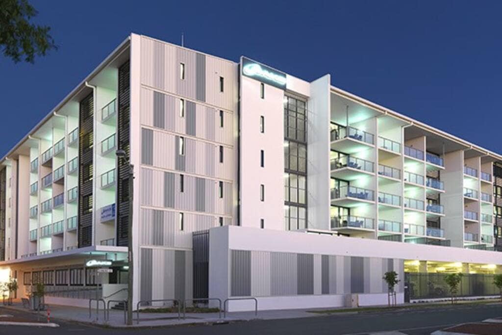 The modern apartment building features a clean, contemporary facade with alternating panels of light and dark materials. Balconies are visible on the upper levels, providing a glimpse of outdoor spaces. The inviting entrance is well-lit, creating a welcoming atmosphere in the evening.