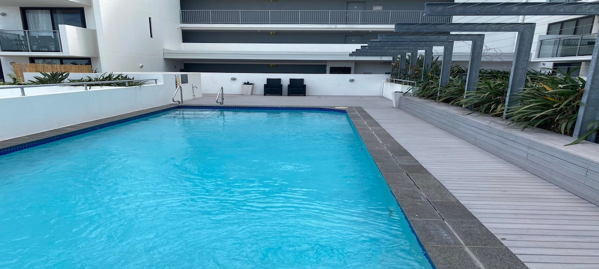 An outdoor pool is surrounded by a wooden deck, featuring clear blue water. Lounge chairs are positioned near the pool, with greenery lining the perimeter. The tranquil setting is complemented by the building's modern architecture visible in the background.