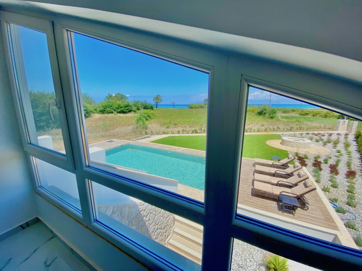 A view from large windows highlights the private pool surrounded by comfortable sunbeds and lush greenery. The tranquil sea is visible in the distance, complementing the serene outdoor space that invites relaxation and enjoyment of the natural surroundings.
