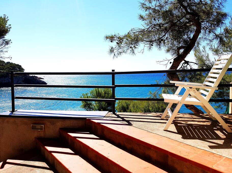 A sunlit terrace features a set of terracotta steps leading down to vibrant blue sea views, bordered by a railing. A white lounge chair is positioned on the terrace, under the shade of pine trees, providing a serene spot to enjoy the coastal scenery.