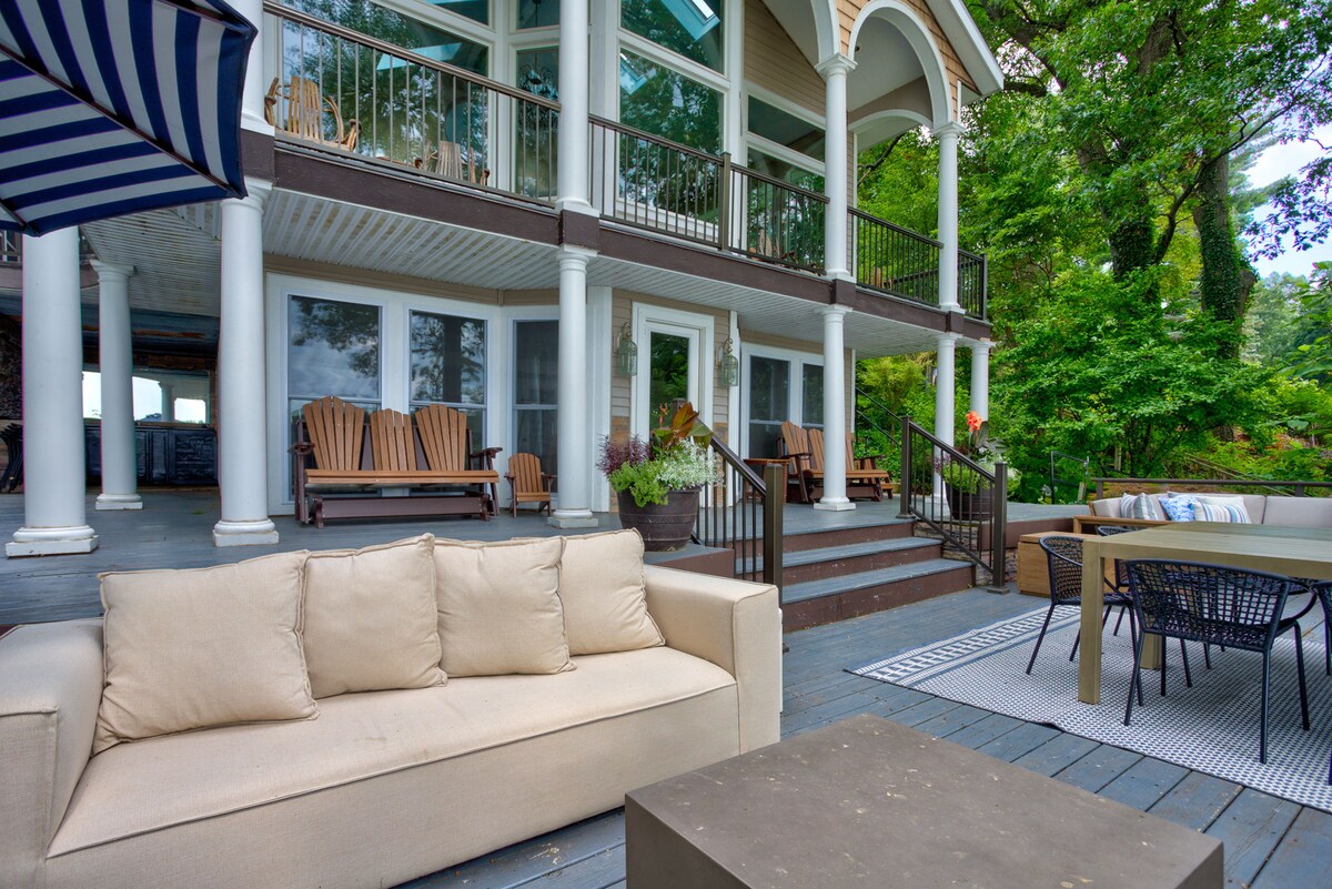 A spacious outdoor deck features elegant seating arrangements, including a comfortable sofa and dining set. The inviting space is framed by lush greenery and large glass windows from the house, providing a seamless connection to nature.