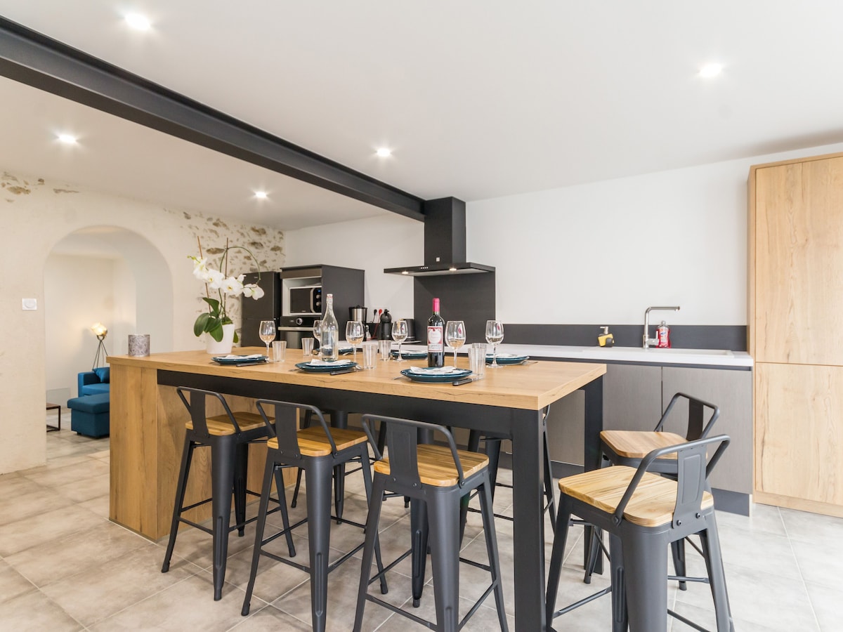 A spacious kitchen area features a large wooden dining table surrounded by black metal chairs. Glasses and place settings are neatly arranged on the table. Sleek cabinetry and modern appliances create a functional cooking space, with soft lighting illuminating the room.