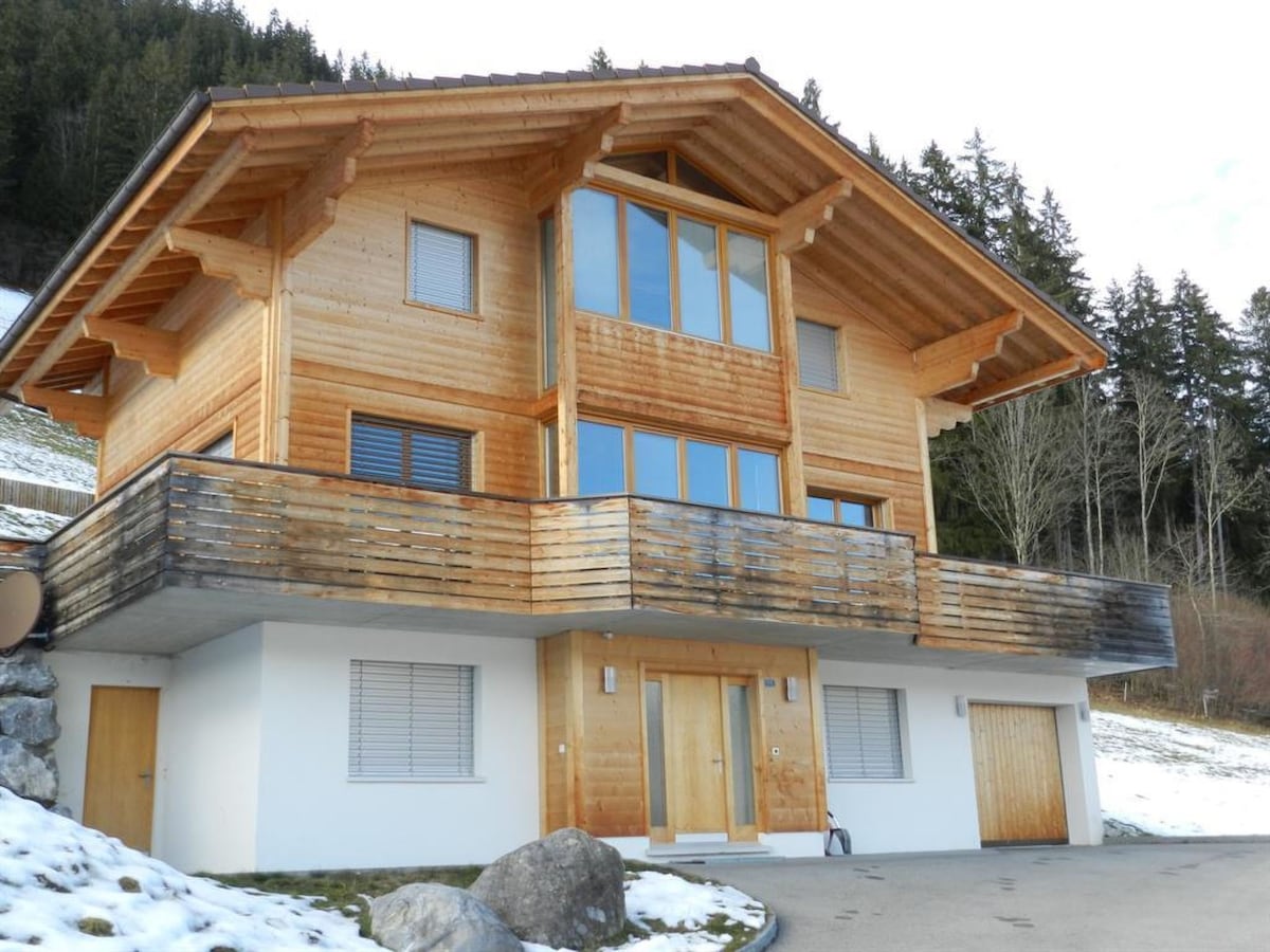 The exterior of a chalet is depicted, featuring a sturdy wooden structure with a spacious balcony and large glass windows. The two-story building is set against a backdrop of snow-covered ground and trees, highlighting its natural surroundings.