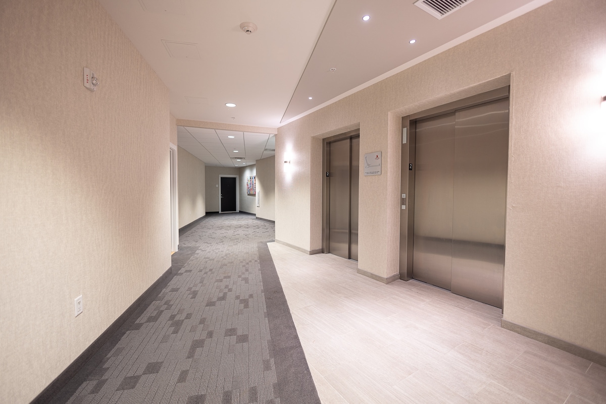 A well-lit hallway is shown, featuring smooth tile flooring and neutral-colored walls. Two elevators are positioned on the right side, with ambient lighting along the ceiling enhancing the spacious feel of the area. The layout is clear, promoting easy navigation.