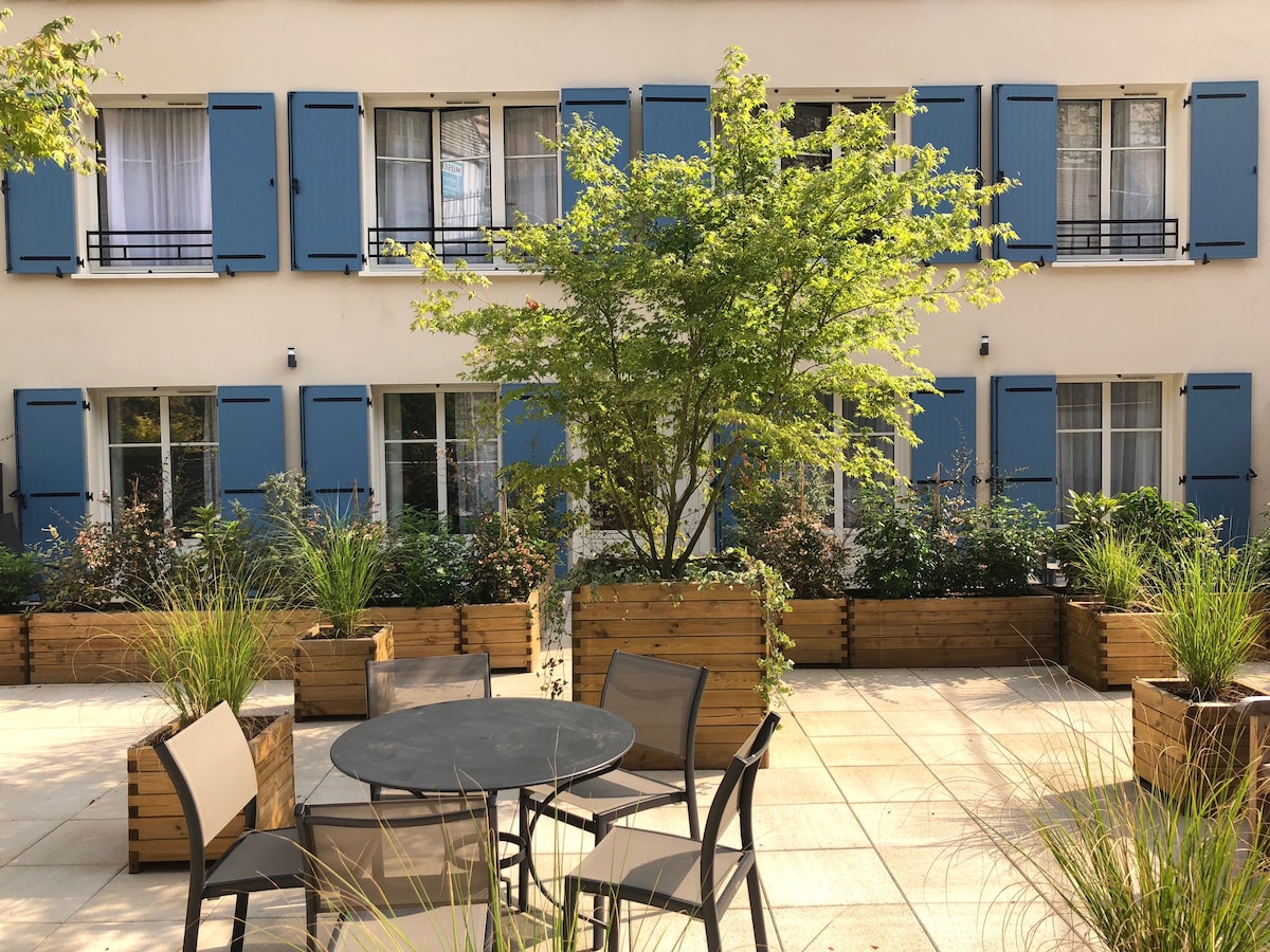 A serene outdoor space features a circular table surrounded by four chairs. Neatly arranged planters filled with greenery line the area, complementing the blue-shuttered windows of the adjacent building. Soft sunlight enhances the inviting atmosphere of the courtyard.