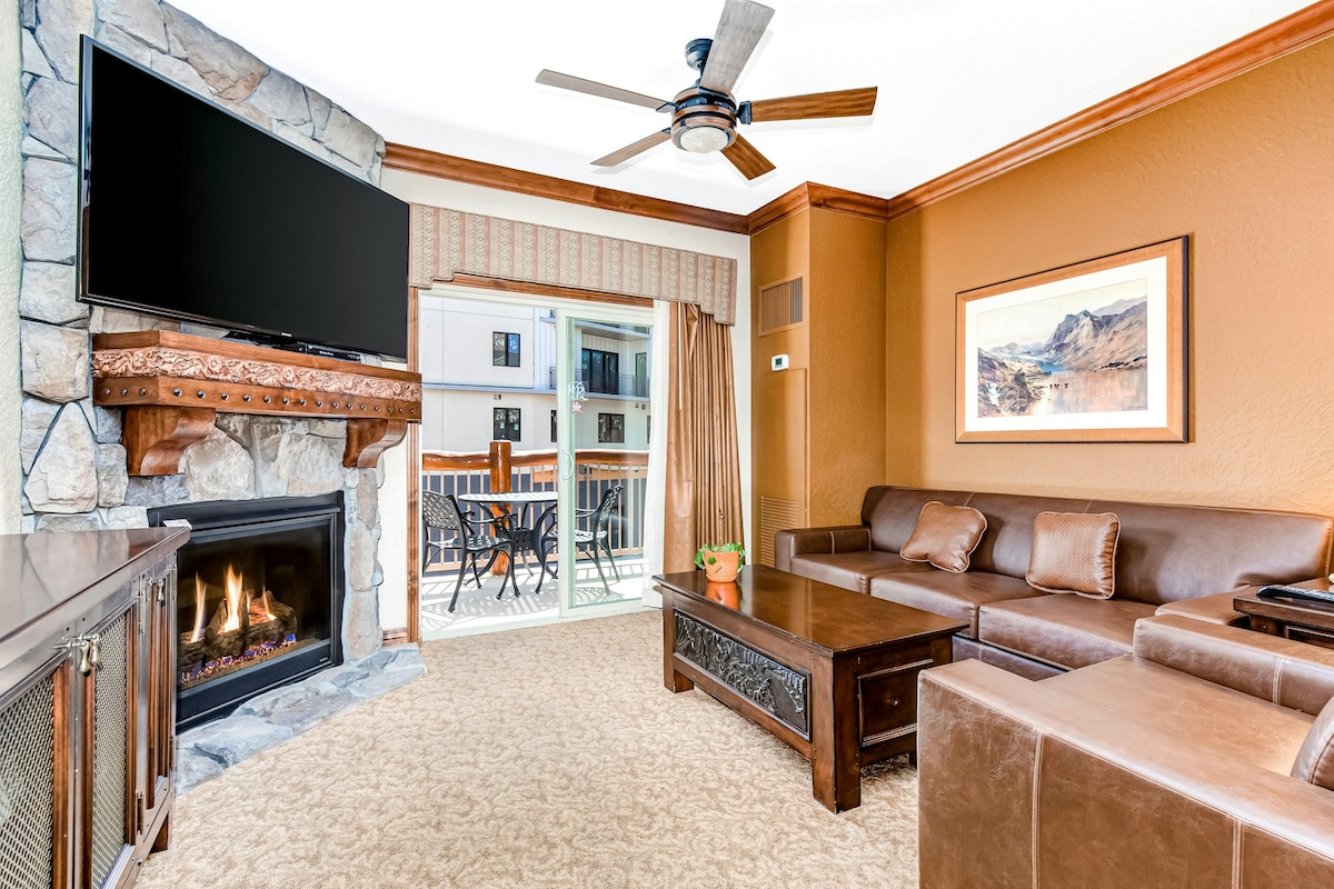 A spacious living area features a stone gas fireplace beneath a flat-screen TV. A comfortable sectional sofa and a wooden coffee table complete the seating arrangement. Natural light floods the room from the sliding glass doors leading to a private balcony furnished with a table and chairs.