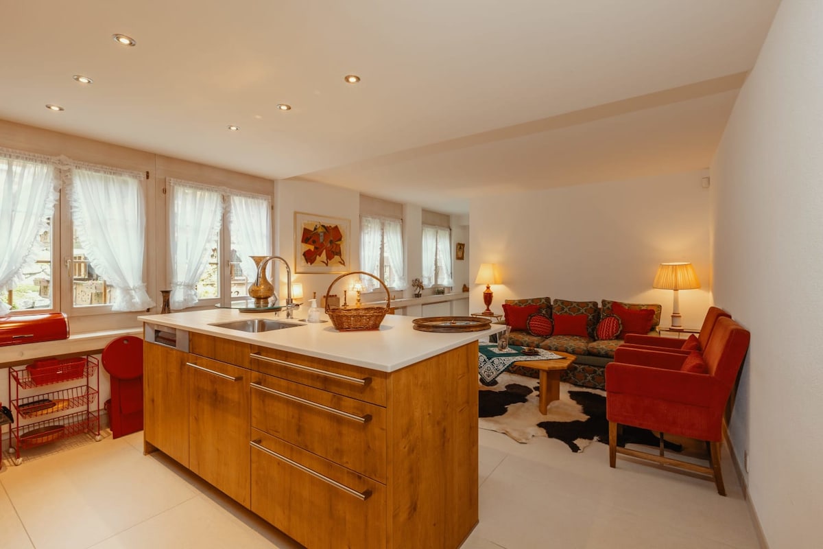 A modern kitchen is highlighted with a large central island featuring a smooth countertop. Natural light filters through large windows adorned with sheer white curtains. A cozy seating area includes a vibrant red sofa and armchair, accompanied by a wooden coffee table and decorative accents.