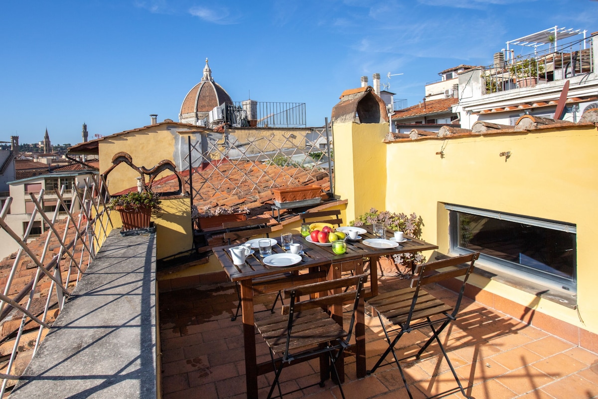 A charming terrace overlooks the cityscape, featuring a dining table set with dishes and fresh fruits. The skyline includes views of iconic rooftops and the dome of the Cathedral of Santa Maria del Fiore. Bright blue skies and a warm ambiance create an inviting outdoor space.