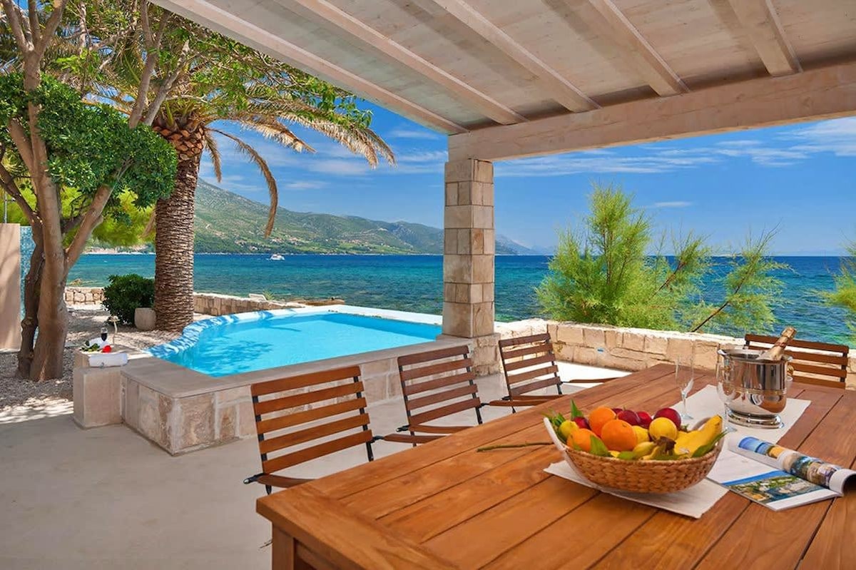 An outdoor terrace is presented with a wooden dining table surrounded by slatted chairs. A basket of fresh fruit rests on the table. In the background, a saltwater pool is visible beside the turquoise sea, framed by palm trees and lush greenery.