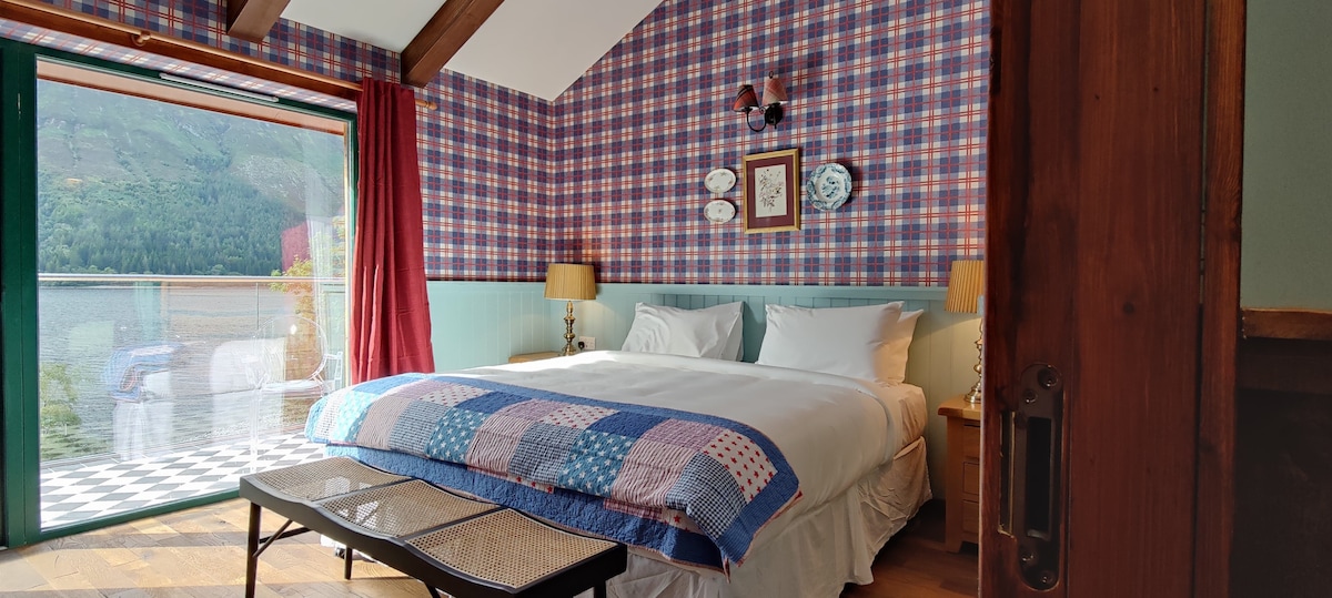 A cozy bedroom features a spacious bed adorned with a patchwork blanket. The walls are decorated with a plaid pattern, and unique wall art adds character. Large glass doors offer access to a view of the lake, complemented by natural light and warm tones.
