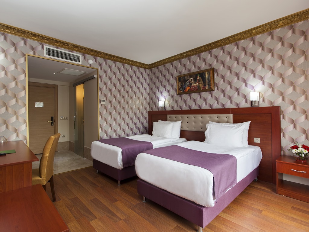 A guest room is shown with two spacious twin beds, each adorned with white linens and purple bedspreads. The walls feature a geometric pattern in soft hues, and a piece of artwork is displayed above the beds. A wooden desk and chair are positioned nearby.