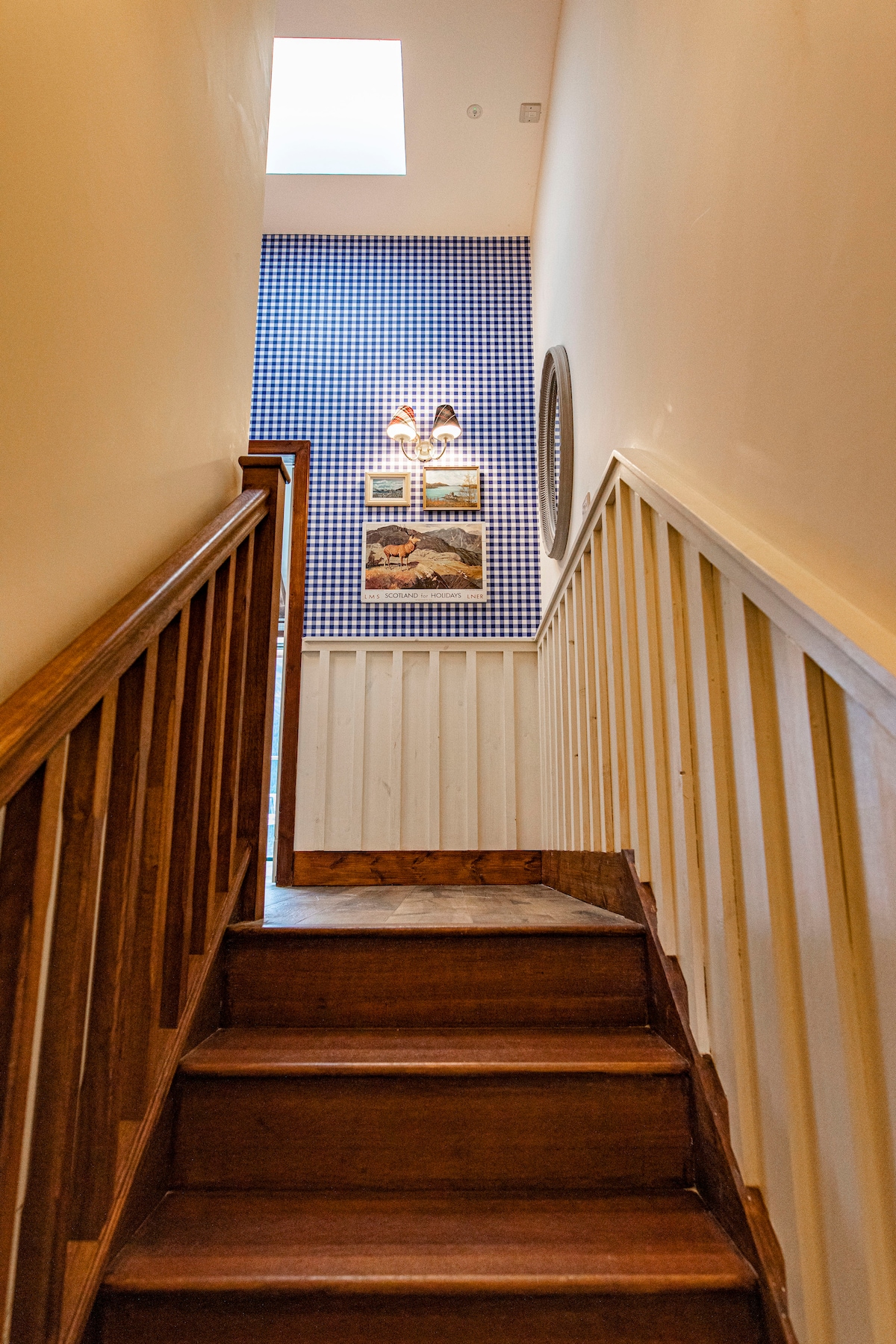 A wooden staircase leads upwards, illuminated by natural light from a skylight. The walls feature a blue and white checkered pattern, complemented by framed artwork. A crisp, clean aesthetic is presented throughout the stairway.