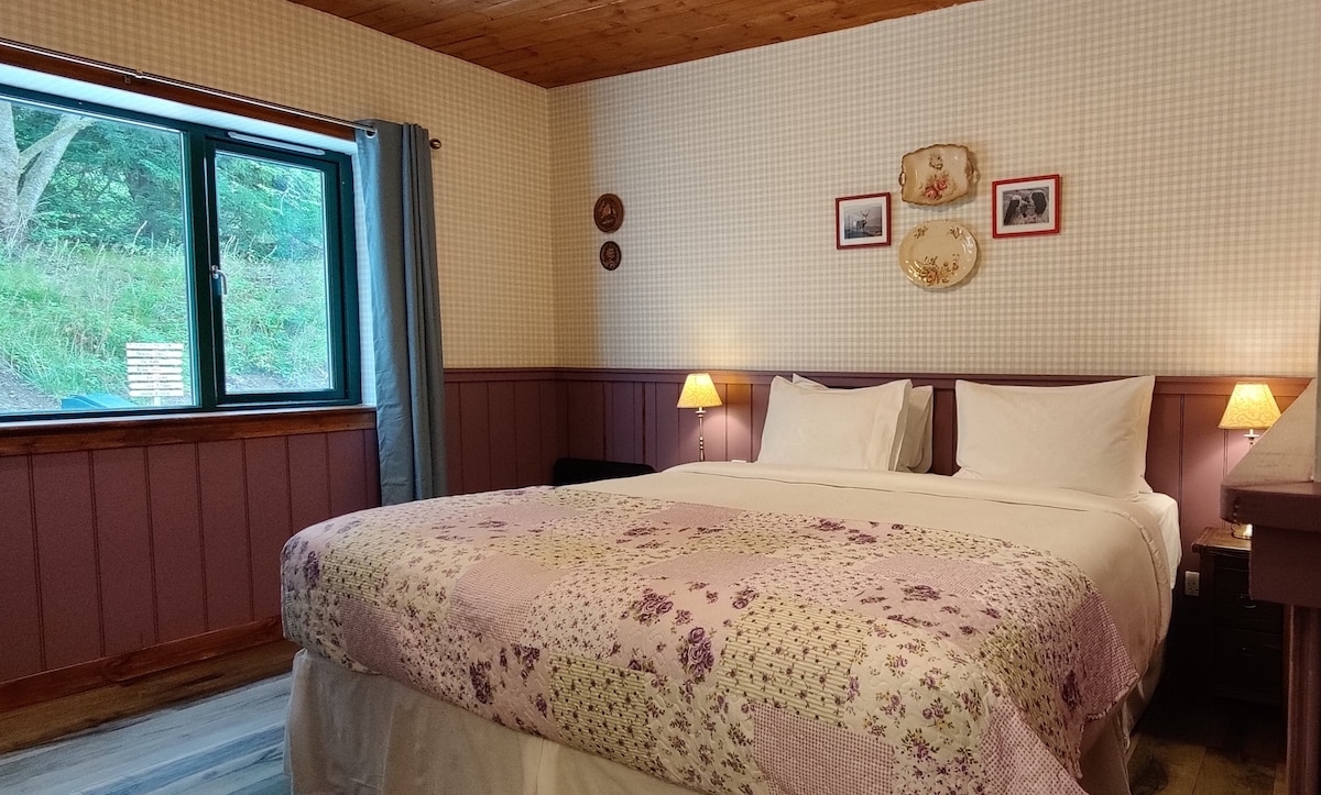 A spacious bedroom is furnished with a large bed dressed in floral bedding. Soft lighting from bedside lamps creates a gentle ambiance. A window with a view of greenery brings natural light into the room, while decorative wall plates and framed photos add character.