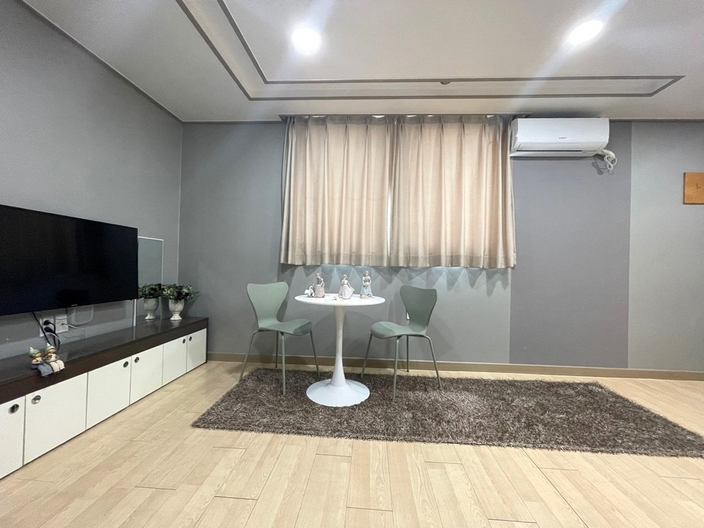 A simple dining area features a round table paired with two green chairs. A television is mounted on the wall opposite the table, with a dark wooden cabinet beneath. Natural light enters through a large window covered by light, sheer curtains, enhancing the serene ambiance.