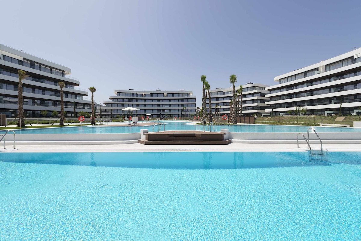 A series of modern buildings are arranged around a large, inviting swimming pool area. The pool features an extensive shallow section, enhanced by clear blue water under a bright sky. Palms and lounge chairs are positioned near the water, creating a relaxing environment.