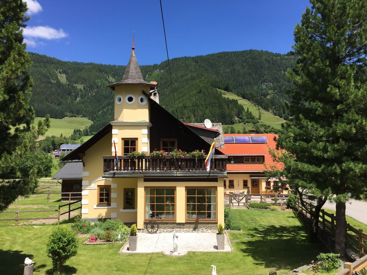 The house features a distinct architectural design with a pointed turret and multiple balconies. Surrounded by manicured gardens and tall trees, the property showcases a serene mountain backdrop under a clear blue sky.