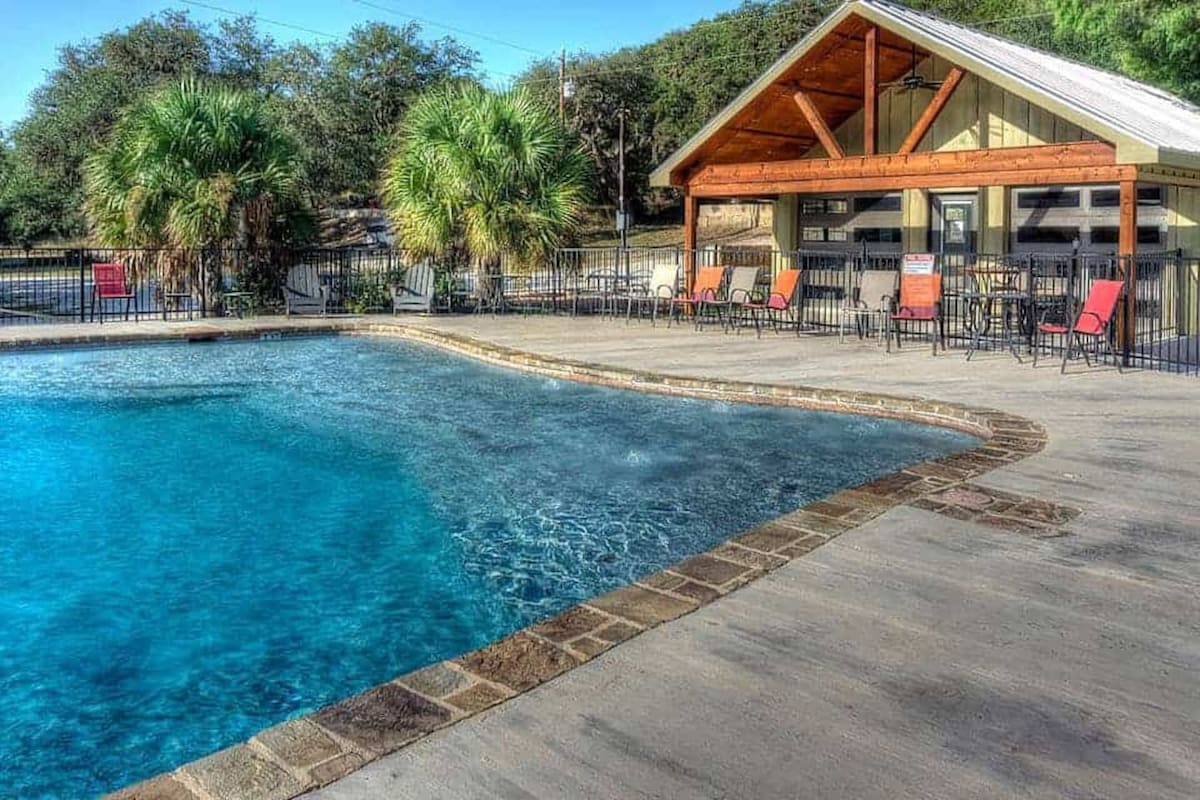 A sparkling swimming pool is surrounded by a spacious patio area, featuring lounge chairs and a covered cabana with large windows. Lush palm trees provide a tropical feel, and the clear water reflects the sunlight, inviting relaxation and enjoyment.