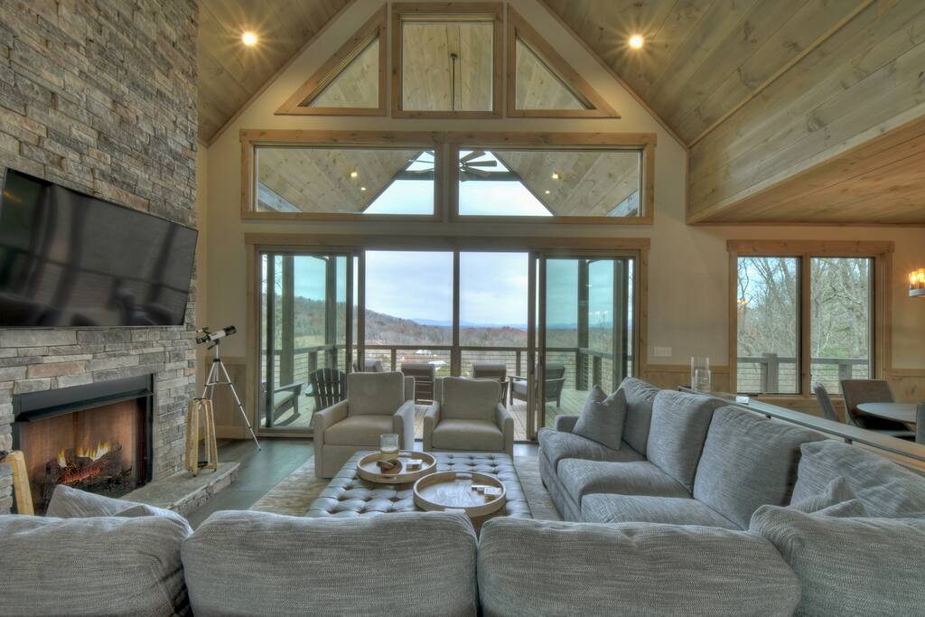An open living room is showcased with a large, comfortable sectional sofa facing a gas fireplace and a wall-mounted TV. Expansive glass doors provide access to a deck with mountain views, while natural light fills the space through high ceilings and large windows.