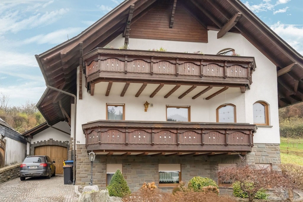 A charming multi-level house features a traditional wooden balcony along the upper floor, adorned with ornamental cutouts. The exterior is complemented by a stone facade and a well-maintained driveway with a parked car. Greenery surrounds the property, enhancing the natural setting.