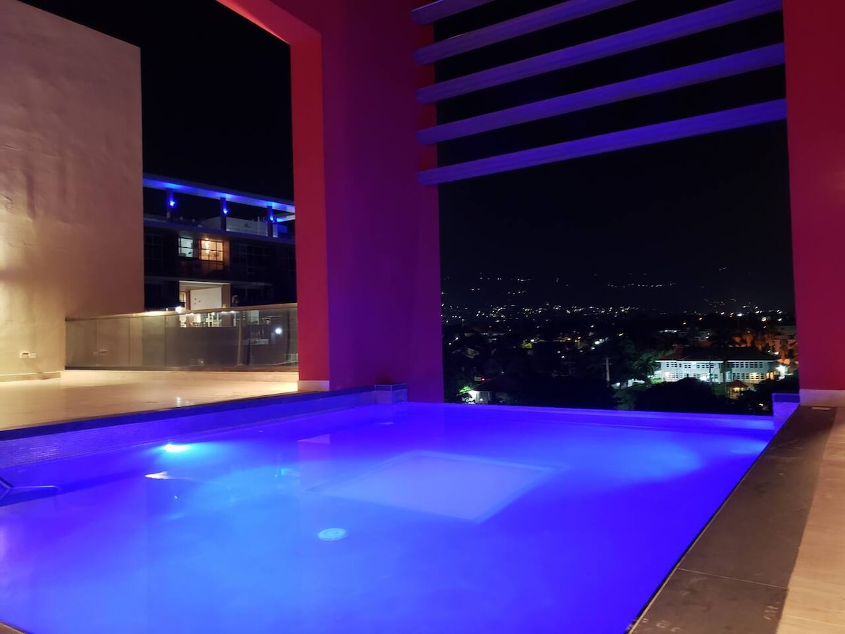 The rooftop pool is illuminated in soft blue light, creating a calming atmosphere. A modern structure with open archways frames views of the surrounding city at night, while the surrounding area showcases vibrant hues reflecting off water. The pool features a sleek design with tranquil edges.