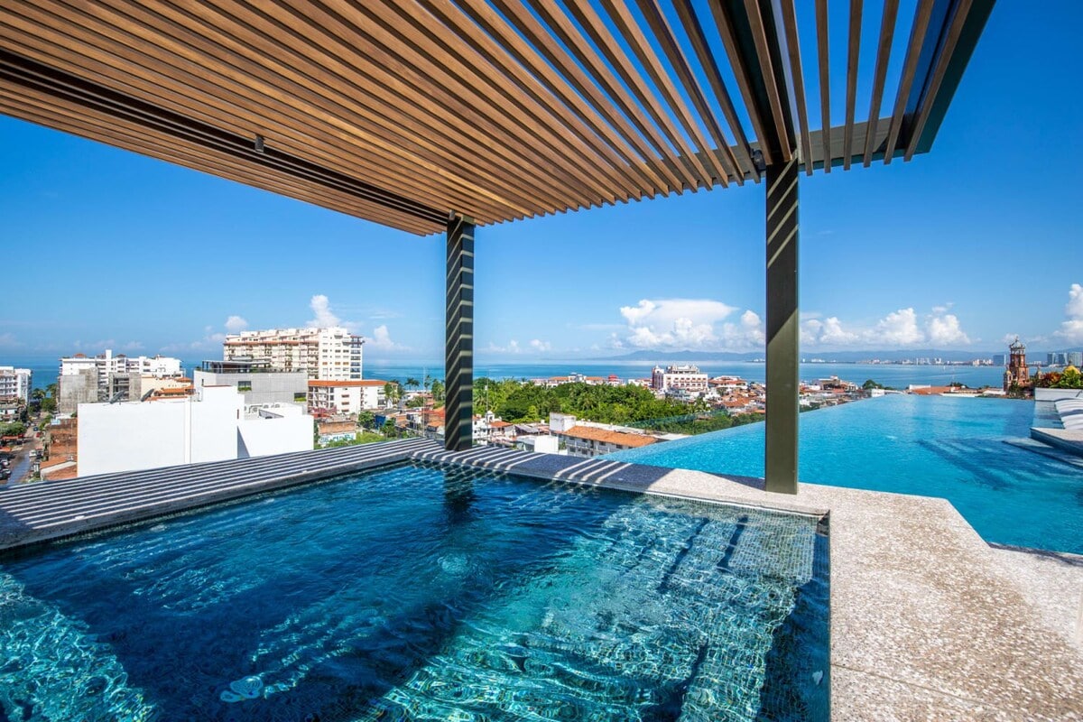 An infinity pool reveals stunning views of the city and coastline, framed by a wooden slatted canopy above. The clear blue water reflects the bright sky, creating a serene and inviting atmosphere for relaxation.